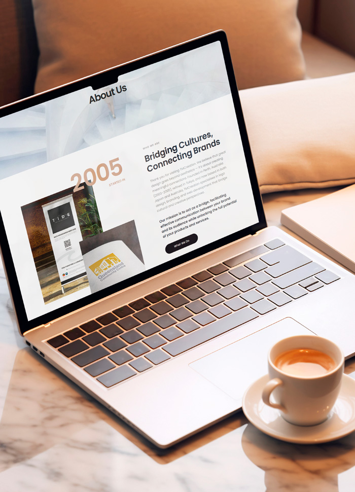 大理石のテーブルに置かれたコーヒーと、TMCreationのウェブサイト（会社概要ページ）を表示したノートパソコン。日本とオーストラリアを拠点にするデザイナーの、洗練された制作ライフスタイルを表現しています。